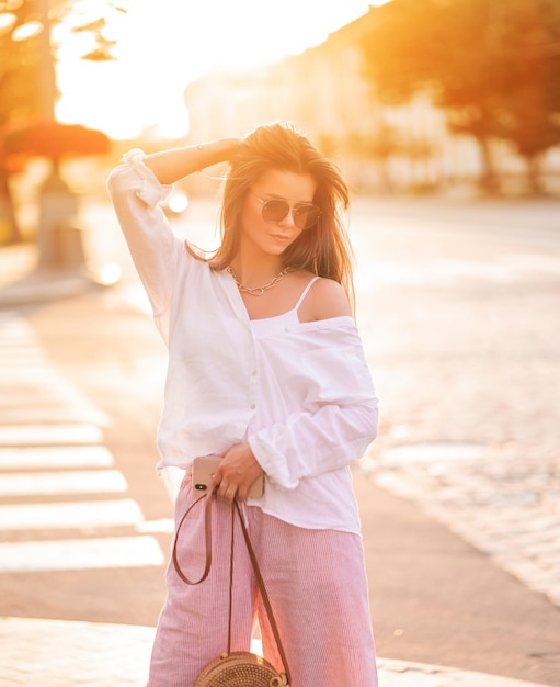 Fille aux cheveux longs dans des verres avec téléphone café et sac se promène dans la ville au coucher du soleil