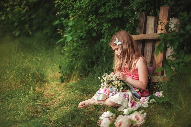 une fille aux cheveux longs est assise sur l'herbe près de la clôture pieds nus et cueille des marguerites