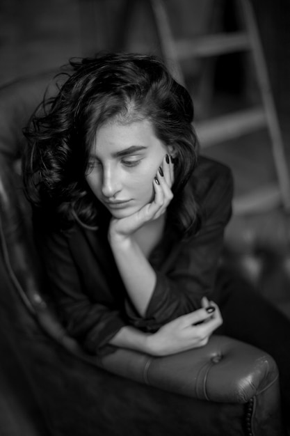 Fille aux cheveux noirs bouclés et maquillage naturel assis sur une chaise en cuir foncé photo noir et blanc