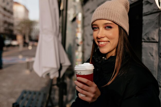 Une fille aux cheveux noirs inspirée de boire du café dans la rue avec un grand sourire