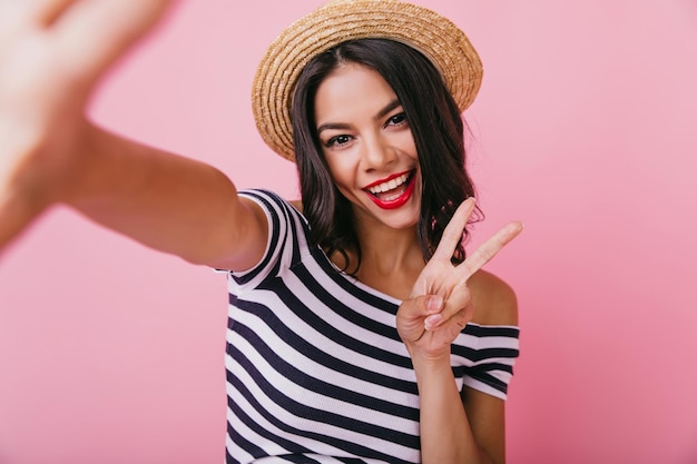 Fille aux cheveux noirs inspirée en riant tout en prenant une photo d'elle-même Belle femme latine débonnaire au chapeau posant sur fond pastel