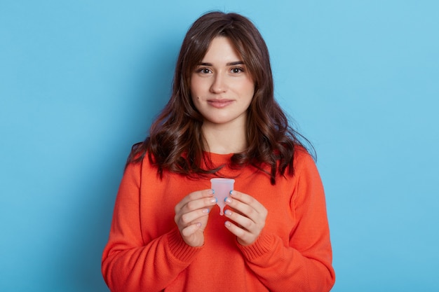 Fille aux cheveux noirs à la recherche agréable avec une expression calme tenant une coupe menstruelle, regardant directement la caméra, isolée sur un mur bleu.