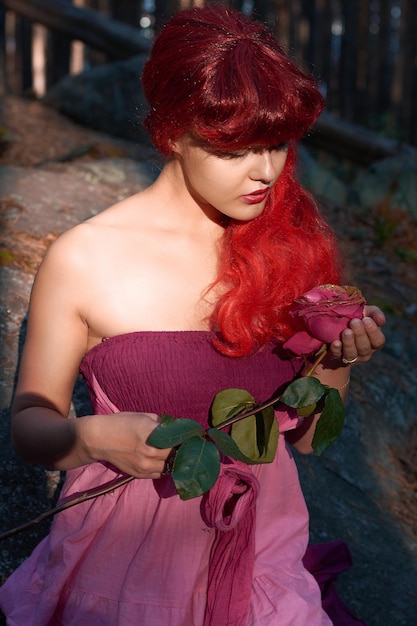 Une fille aux cheveux rouges et une robe avec une rose dans les mains se repose dans une fabuleuse forêt