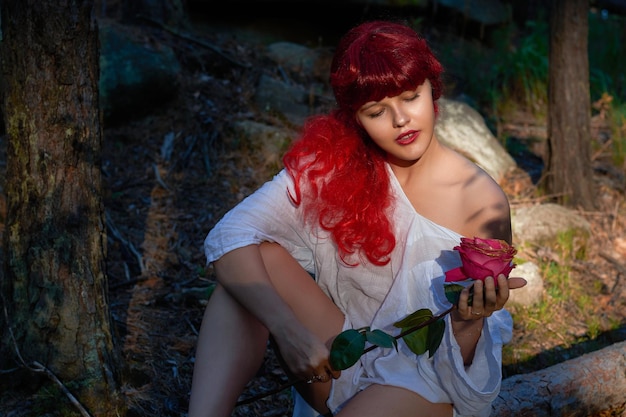 Une fille aux cheveux roux et une chemise blanche avec une rose dans les mains se repose dans une fabuleuse forêt