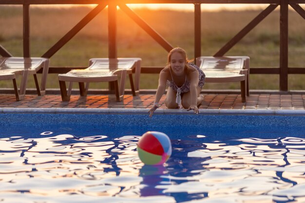 Une fille aux cheveux tressés en chignon en costume lumineux joue au bord de la piscine avec un ballon sur fond de soleil d'été