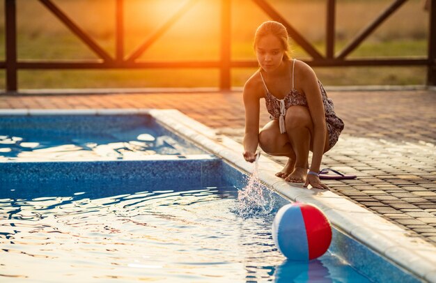 Une fille aux cheveux tressés en chignon en costume lumineux joue au bord de la piscine avec un ballon sur fond de soleil d'été