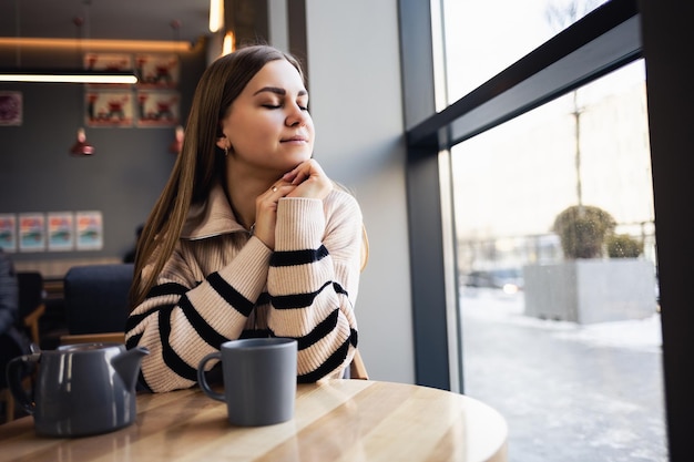 Une fille aux yeux bruns dans un pull en laine s'appuie sur une table dans un café et regarde par la fenêtre Une jeune femme est venue boire une tasse de thé chaud parfumé dans un café