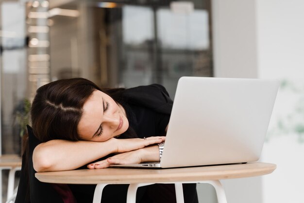 Une fille aux yeux fermés fait une pause au travail et dort et se détend sur un ordinateur portable sur le lieu de travail en raison d'heures supplémentaires