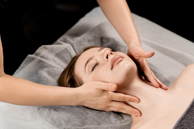 Une fille aux yeux fermés se détend sur un massage du visage et du cou dans un spa Le masseur fait un massage du visage pour une jeune femme dans un spa