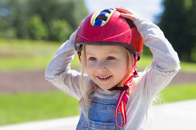 Fille de bambin heureux portant un casque en plastique rouge à l'extérieur. Portrait de jolie fille de 4 ans tenant un casque sur la tête avec les mains