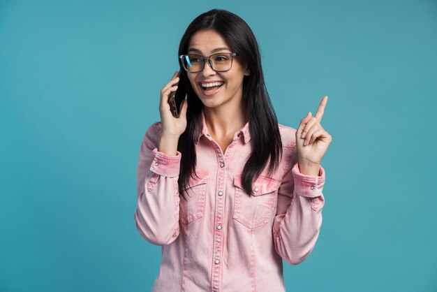 Une Fille Bavarde. Portrait D'une Jeune Femme Sortie Debout, Faisant Des Gestes Et Passant Un Appel Sur Son Smartphone Avec Un Visage étonné Et Parlant Avec Un Ami. Intérieur, Isolé, Tourné En Studio, Fond Bleu