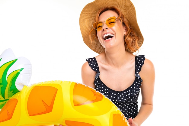 Fille avec un beau sourire vêtu d'un maillot de bain rétro et un chapeau de paille détient un cercle de natation en forme d'ananas sur un mur blanc