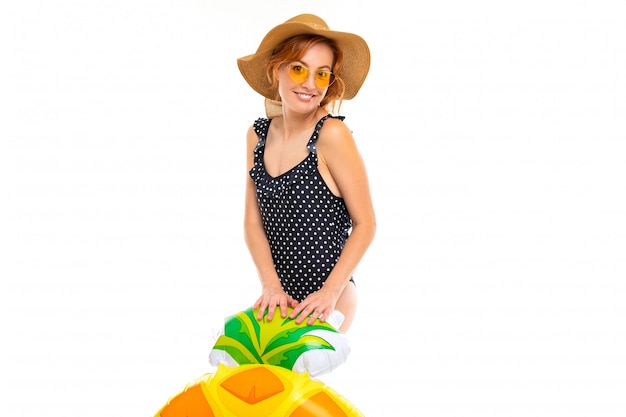 Fille avec un beau sourire, vêtue d'un maillot de bain rétro noir et d'un chapeau de paille, tient un cercle de natation en forme d'ananas sur un mur blanc