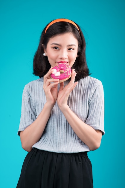 Fille de beauté asiatique tenant un beignet rose. Femme joyeuse rétro avec des bonbons, dessert debout sur fond bleu.