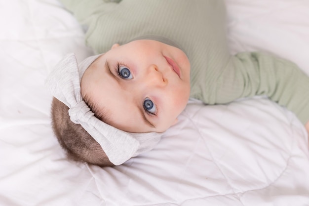 Une fille avec de beaux grands yeux frotte le bébé à la maison sur le lit dans un body en coton sur du linge de lit blanc