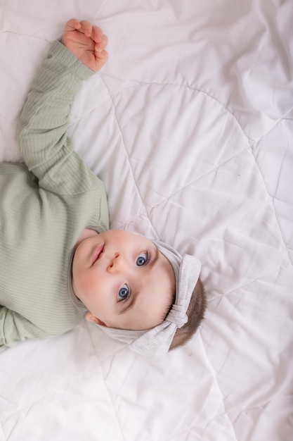 Une fille avec de beaux grands yeux frotte le bébé à la maison sur le lit dans un body en coton sur du linge de lit blanc
