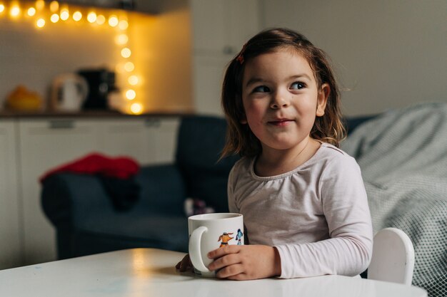 Fille de bébé caucasien, boire du cacao dans une tasse à la maison. Lumières de bokeh de Noël. Photo de haute qualité