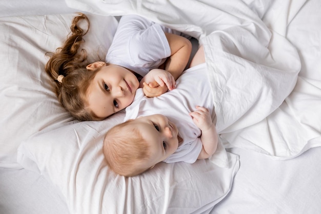 Fille et bébé en vêtements blancs sont allongés dans le lit sur du linge de lit blanc frère et soeur se prélassent au lit