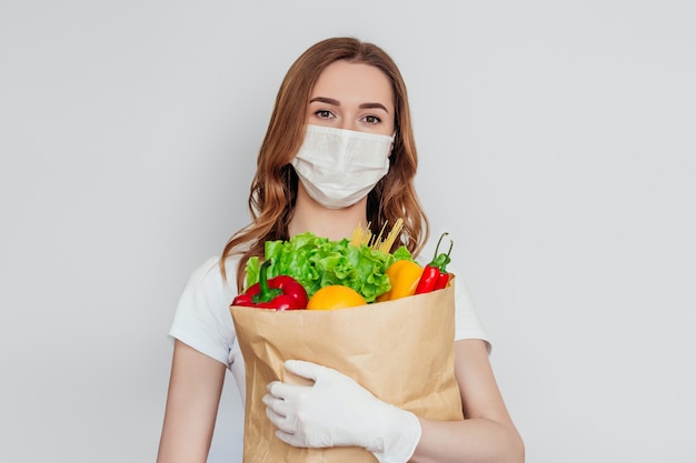 Fille bénévole courrier portant un masque médical est titulaire d'un sac en papier avec des produits, légumes, poivrons, salade isolé sur blanc, espace gris