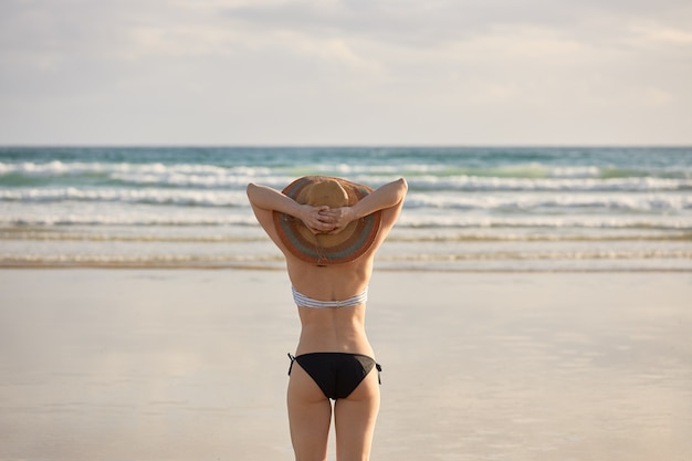 Fille en bikini avec chapeau de paille sur le rivage d'une plage tenant son chapeau de paille.