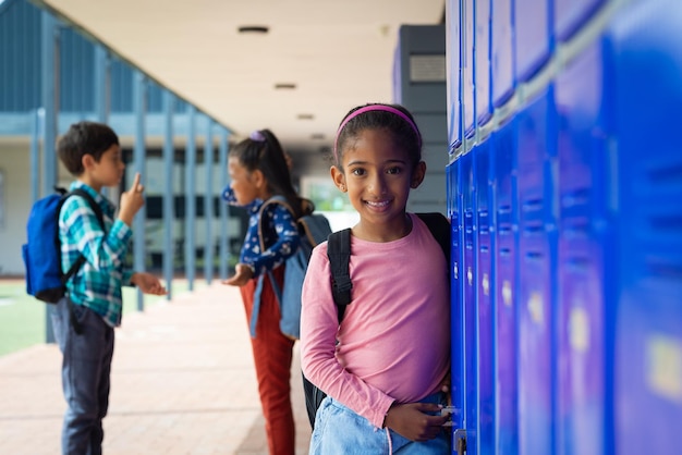 Une fille biraciale avec un sac à dos rose sourit près des casiers bleus de l'école ses cheveux noirs attachés en arrière