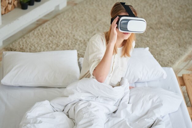 La fille en blanc assise sur le lit dans le casque de réalité virtuelle