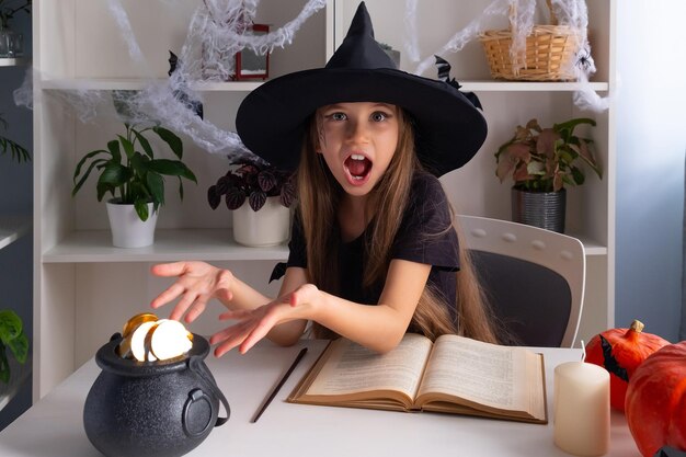 Une fille blonde de 7 à 8 ans en costume de sorcière célèbre Halloween avec un livre de sorcière et un pot de bonbons en or