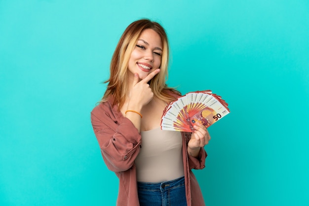 Fille blonde adolescente prenant beaucoup d'euros sur fond bleu isolé souriant