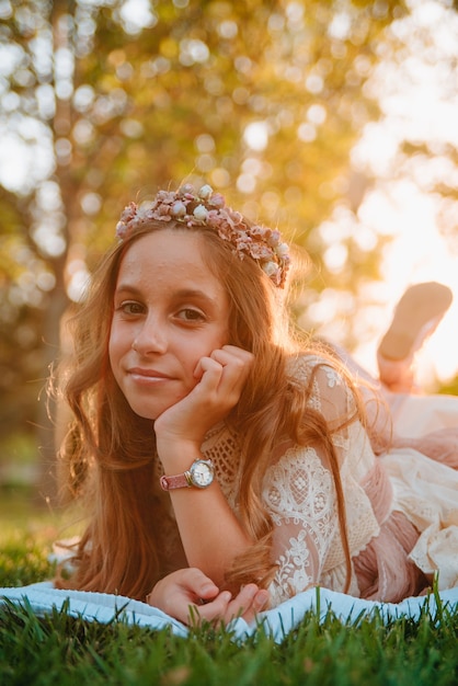 Fille blonde aux cheveux bouclés vêtue d'une robe de communion allongée sur l'herbe pour des photos