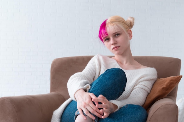 Une fille blonde aux cheveux roses portait un chandail blanc confortable et un jean est assis sur une chaise à la maison