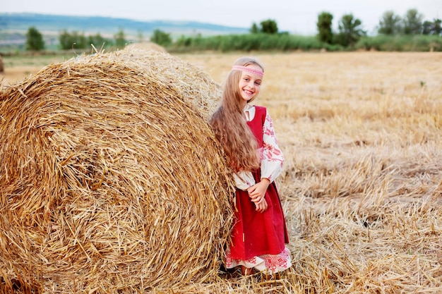 une fille blonde aux grands yeux bruns dans un costume slave se tient près d'une botte de foin