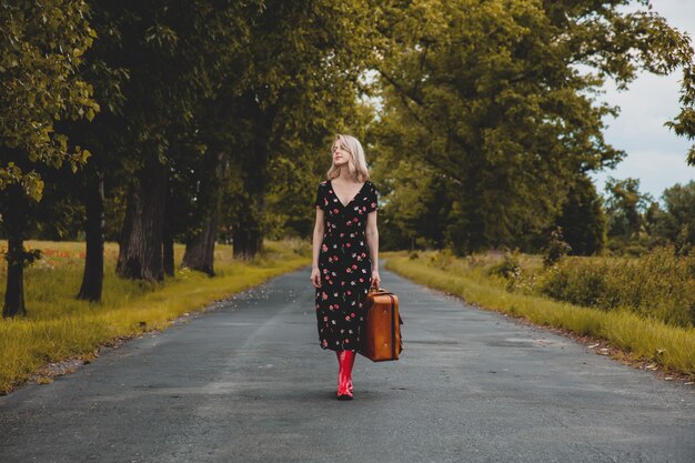 Fille blonde en bottes rouges avec suicase à la route de campagne