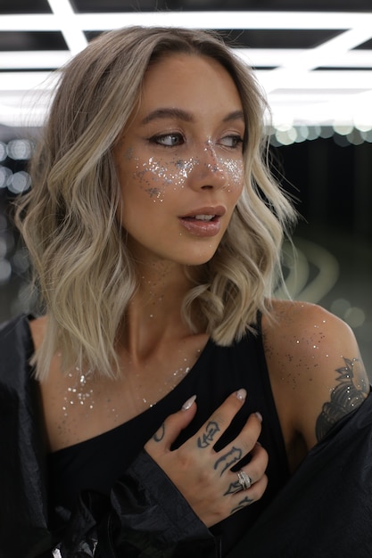 Une fille blonde brillante avec des paillettes sur le visage, le cou et les épaules se tient à l'intérieur avec une lumière vive et un look attrayant sur le côté photo de haute qualité