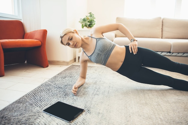 Fille blonde caucasienne faisant des exercices de fitness à domicile et portant des vêtements de sport utilise une tablette sur le sol