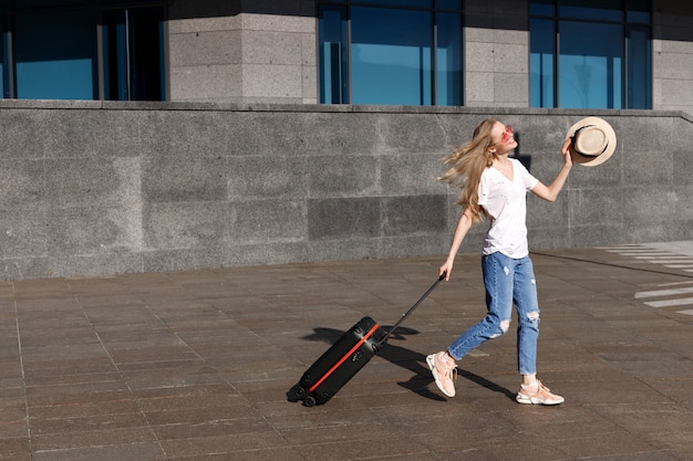 Une fille blonde avec un chapeau de paille et une valise pour voyager fait le tour de la ville en été en globe-trotter