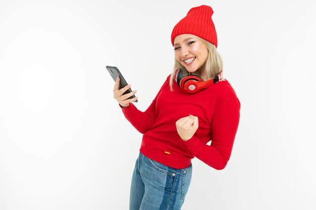 Fille blonde charismatique énergique avec un smartphone et un casque dansant sur un fond blanc.