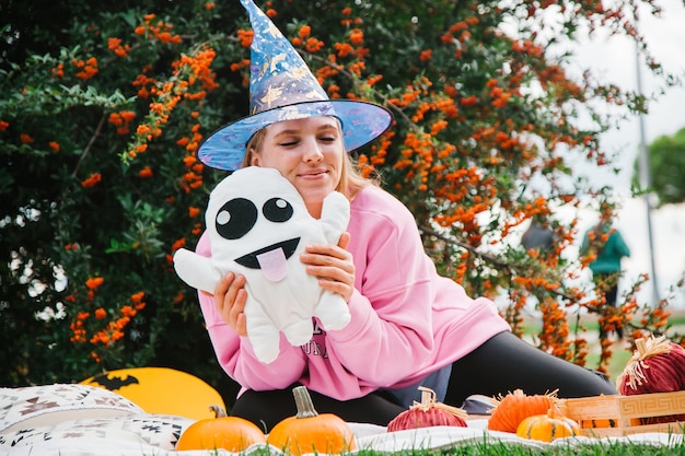 Fille blonde sur la couverture dans le parc sur la pelouse avec un fantôme et des citrouilles symboles d'Halloween