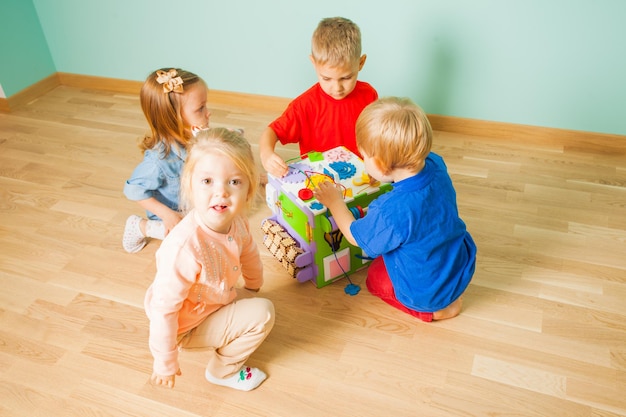 Fille blonde curieuse regardant la caméra, assise sur un sol près de ses amis, qui jouent avec un cube occupé. Développer des jouets pour la maternelle ou la garderie