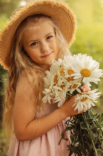 une fille blonde dans un chapeau de paille dans une robe en lin rose tient un bouquet de marguerites dans ses mains