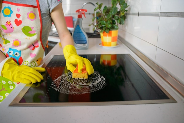 Fille blonde dans les gants jaunes avec une éponge essuyant la cuisinière à induction électrique en céramique haut de gamme grand angle Espace de copie