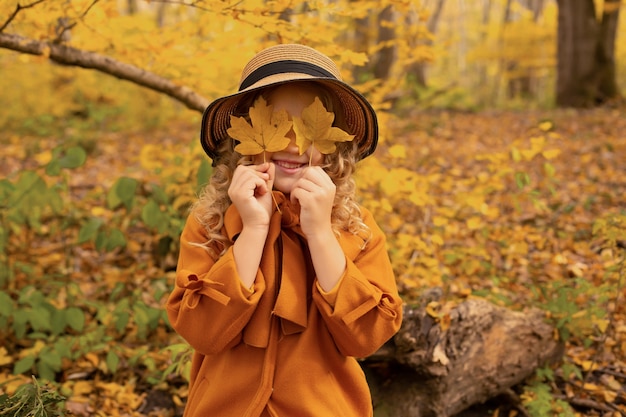 fille blonde dans un manteau couleur moutarde dans un chapeau dans une forêt urbaine d'automne avec des feuilles dans ses mains