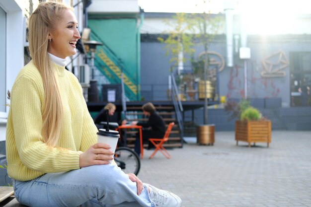 Une fille blonde dans un pull jaune et un jean est assise sur un banc avec une tasse de café à emporter la fille l