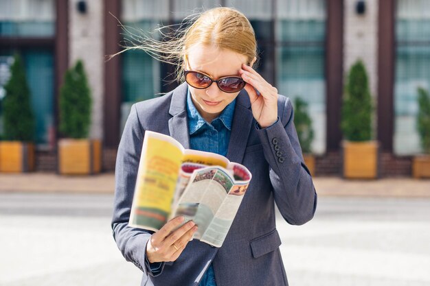 Une fille blonde dans une veste grise et des lunettes brunes lit la presse fraîche avec surprise