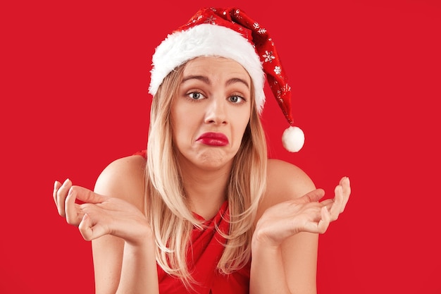 Fille blonde du père noël confuse avec une expression de visage douteuse et indécise au nouvel an et noël isolée sur fond rouge, espace de copie vide