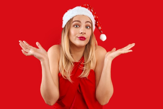 Fille blonde du père noël confuse avec une expression de visage douteuse et indécise au nouvel an et noël isolée sur fond rouge, espace de copie vide