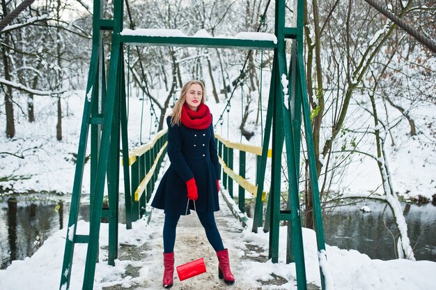 Fille blonde en écharpe rouge et manteau marchant sur le pont au parc le jour d'hiver.