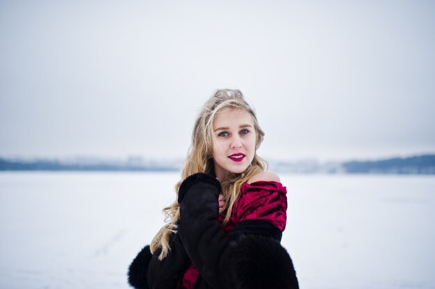 Fille blonde élégance en manteau de fourrure et robe de soirée rouge posant au jour de neige en hiver.