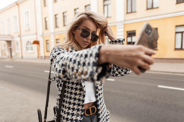Fille blonde élégante dans des vêtements à la mode avec des lunettes de manteau et un sac à main se promène dans la ville et prend une photo de selfie