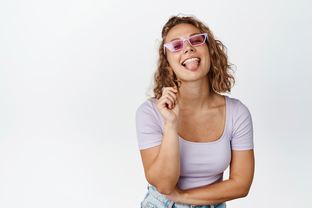 Fille blonde élégante à lunettes de soleil, souriant et riant, debout en t-shirt blanc