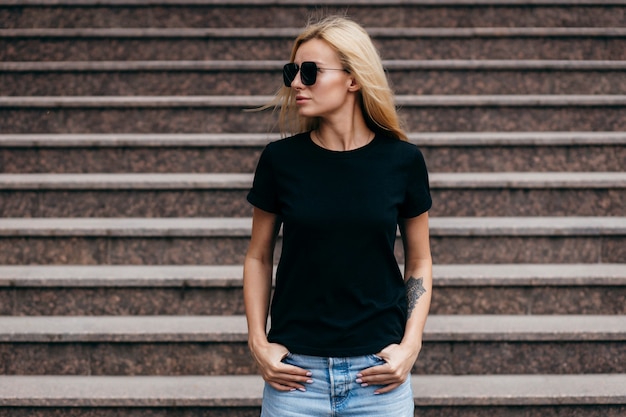 Fille blonde élégante portant un t-shirt noir et des lunettes posant contre la rue